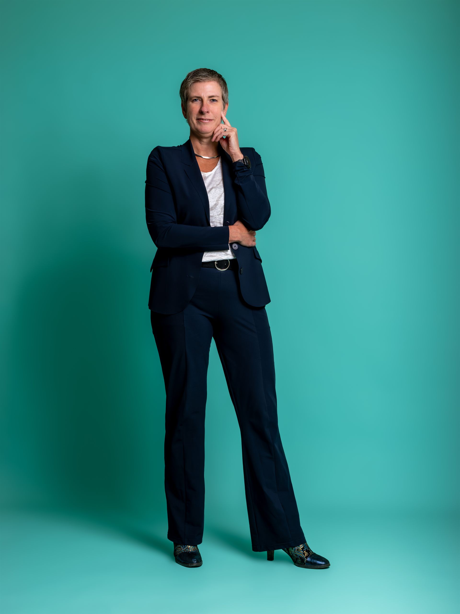 Zakelijke headshot van een glimlachende vrouw in een donkerblauw pak tegen een turquoise achtergrond.